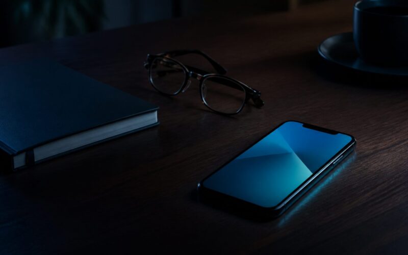 Smartphone glowing beside a notebook and reading glasses on a dark desk